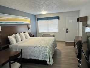 A hotel room with a bed, a desk, a chair, a window with blinds, a door, and a wall-mounted air conditioning unit. - HomeTowne Studios by Red Roof Georgetown, KY (Georgetown)
