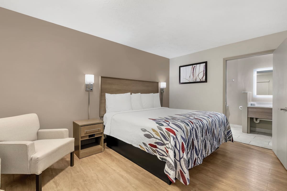A hotel room with a bed, a nightstand, a chair, a wall-mounted light, and a door leading to a bathroom.