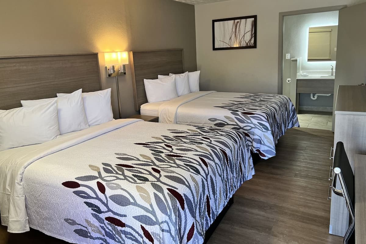 A hotel room with two beds, a headboard, wall-mounted lamps, a framed picture, and a bathroom visible through an open door.