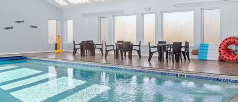 An indoor swimming pool with clear water, surrounded by wooden flooring and a seating area with chairs and tables.