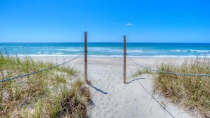 Une plage de sable bordée d’une barrière faite de cordes et de poteaux en bois, qui mène à l’océan.