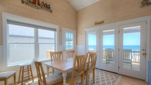 Une pièce avec une table et des chaises en bois, de grandes fenêtres donnant sur l’océan et une porte menant à un balcon.