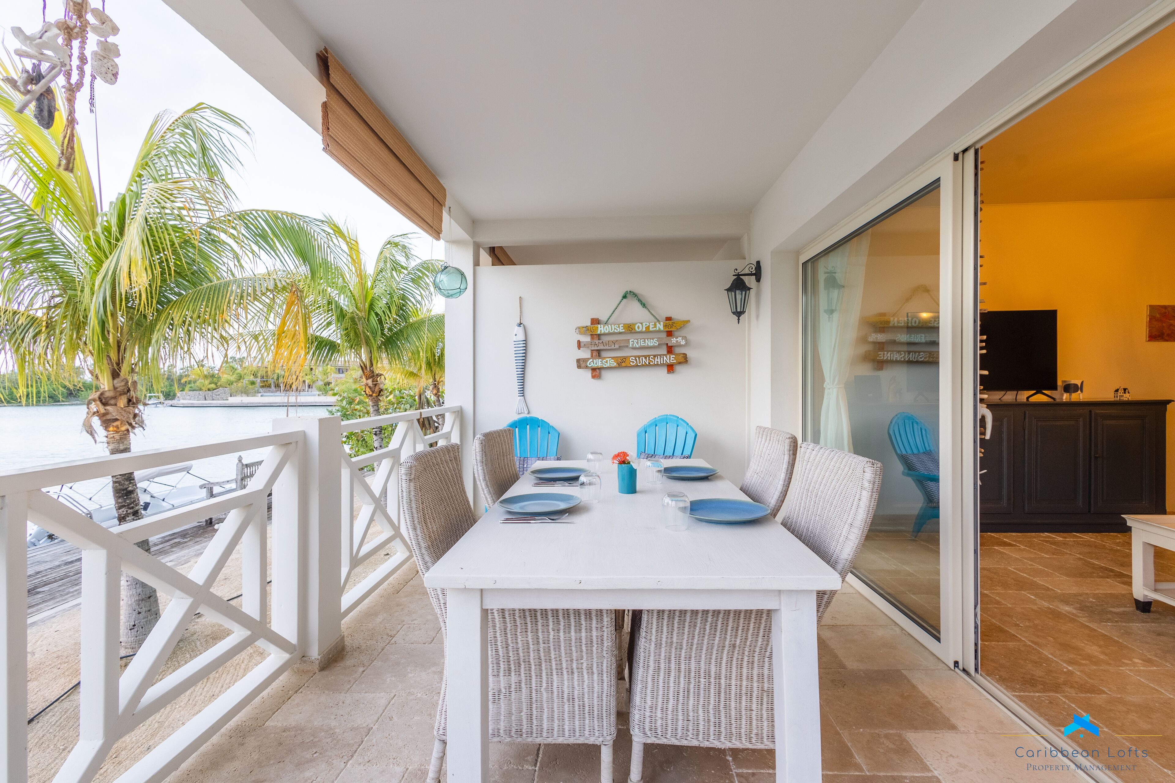 A white dining table with blue plates and wicker chairs, a sign indicating the house is open, and a view of a palm tree and ocean.