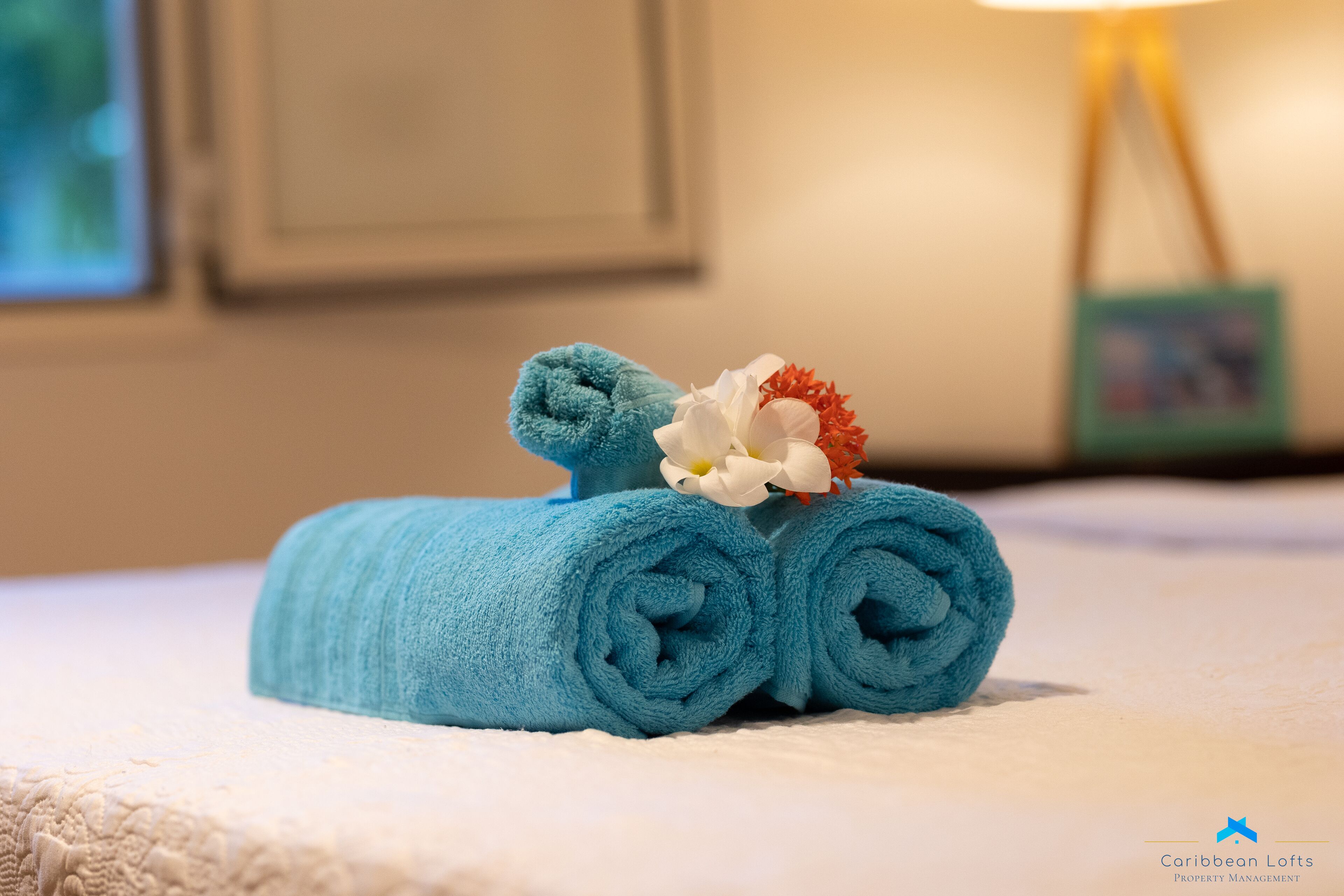 Een netjes opgerolde blauwe handdoek met een witte en rode bloem op een bed.