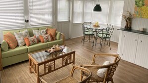 A living room with a green sofa, a glass coffee table, wicker chairs, and a dining area with a round table and chairs. - Coquina Cottage on Vilano Beach (St. Augustine)