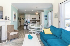 A modern living room with a blue sofa, a wicker chair, a white coffee table, and a kitchen in the background.