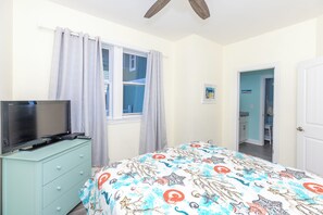 A bedroom with a bed, a TV on a dresser, a window with curtains, and a door leading to another room.