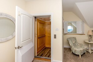 A hotel room with a wooden door, a round mirror, a floral armchair, a small table with a lamp, and a view through a window with shutters.