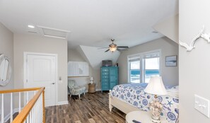 Un dormitorio con cama, cómoda, silla, ventilador de techo y vista al océano a través de amplios ventanales.