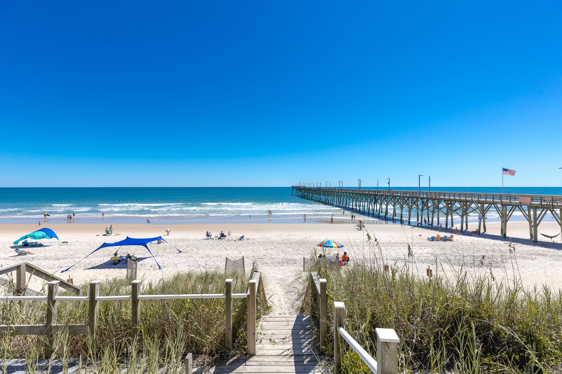 A beach with a wooden pier extending into the ocean, a clear sky, and a sandy shore with people and umbrellas.