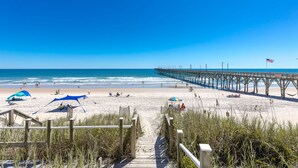 A beach with a wooden pier extending into the ocean, a clear sky, and a sandy shore with people and umbrellas. - Pier Pressure - Amazing Oceanfront 6 BR Half Duplex; Hot Tub and more! Pet (Surf City)
