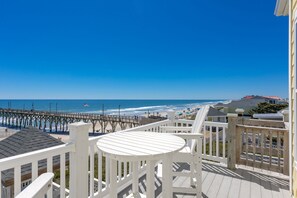 A balcony with a view of the beach and ocean, wooden deck, and a pier in the background.