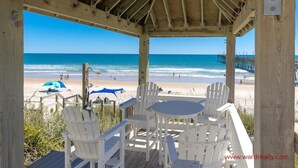 Eine Holzterrasse am Strand mit weißen Stühlen und einem Tisch, mit Blick auf einen Strand und einen Pier in der Ferne.