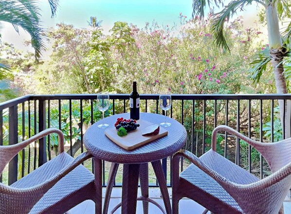 A round table with wicker chairs, a bottle of wine, and a glass on a balcony with a view of lush greenery and trees.