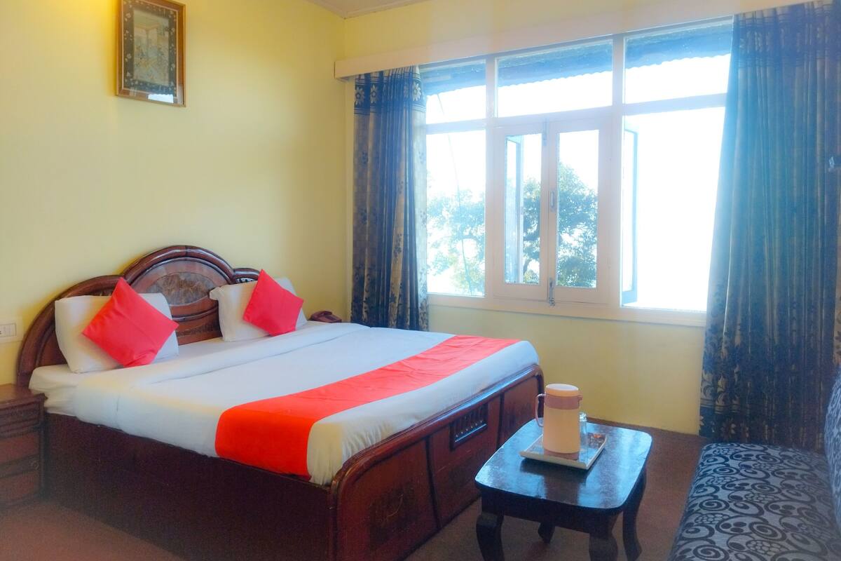A hotel room with a bed, a bedside table with a coffee pot, a window with curtains, and a framed picture on the wall.