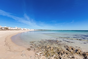 A beach with clear water, sandy shore, and buildings in the background.