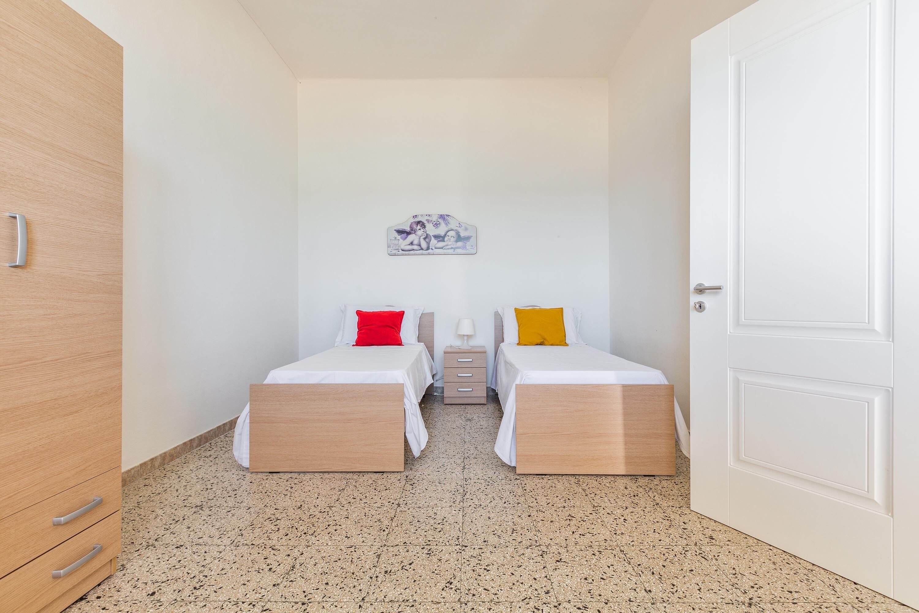 A small, clean room with two single beds, a wooden wardrobe, a small table, and a white door.