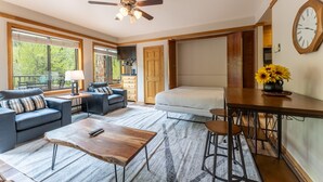 Una habitación amplia con una cama, una mesa de comedor y un sofá. Hay un ventilador de techo, un reloj en la pared y una ventana con persianas.