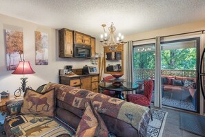 A cozy living room with a sofa, a dining table, a chandelier, and a view of a balcony with outdoor seating.