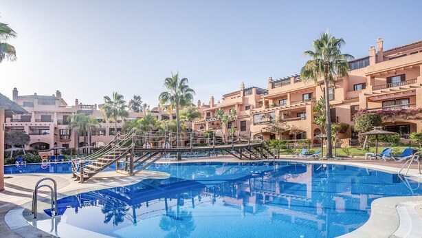 Ein Pool mit Holzterrasse und Treppe, umgeben von Palmen und einem Gebäude mit Balkonen.