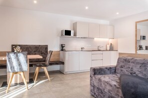 Une cuisine moderne avec des meubles blancs, un four à micro-ondes et une machine à café. On y trouve un coin repas avec une table et des chaises en bois, ainsi qu’un canapé gris.