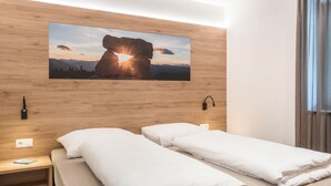 Hotellihuone, jossa on puinen sängynpääty, valkoisilla lakanoiden peitetty sänky, yöpöytä, jossa on kirja, ja seinälle ripustettu kuva kalliojyrkänteestä auringonpaisteessa.