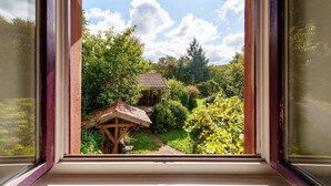 Vista attraverso una finestra su una struttura in legno con tetto in tegole, immersa in un verde rigoglioso e un cielo sereno.
