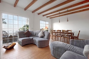 A spacious living room with a large L-shaped grey sofa, a dining area with wooden chairs and a table, and large windows allowing natural light. - Pet-Friendly Vacation Home "Casa Luz" with Sea View, Whirlpool & Wi-Fi (La Luz)