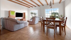A spacious living area with a sofa, TV, dining table, and chairs, wooden beams on the ceiling, and large windows. - Pet-Friendly Vacation Home "Casa Luz" with Sea View, Whirlpool & Wi-Fi (La Luz)