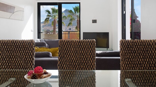 Une pièce avec une table en verre, deux chaises à motifs, un canapé, un téléviseur et une vue sur des palmiers et des montagnes, visible à travers une porte coulissante.