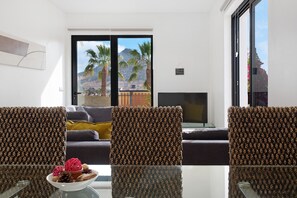 A room with a glass table, two patterned chairs, a sofa, a TV, and a view of palm trees and mountains through a sliding door. - Villa "Recanto Dourado" with Sea View, Private Garden & Wi-Fi (Porto Santo)