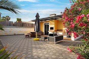Eine Terrasse mit sechseckigen Fliesen, Gartenmöbeln, einer Pergola und einem Strauch mit Blumen.