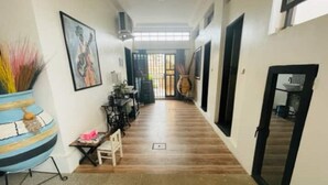 A hallway with a wooden floor, a dining area with chairs and a table, a large vase with plants, and a painting on the wall.