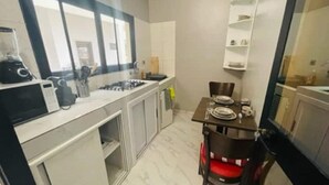 A compact kitchen with a microwave, stove, and sink. There is a dining table with chairs and a small shelf above it.