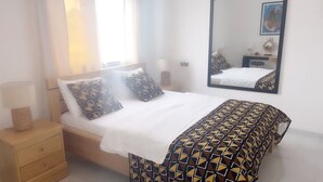A bedroom with a bed, two pillows, a bedside table, a lamp, a mirror, and a dresser.