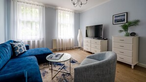 A living room with a blue sofa, a grey armchair, a white dresser, a TV, and a chandelier. - Parkowa 33A2 - Beachfront Apartment with Parking by Lion Apartments Sopot (Sopot)