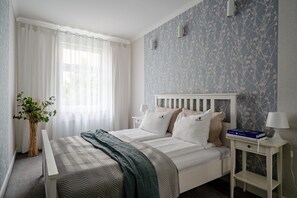 Un dormitorio con una cama, mesitas de noche, una lámpara, una planta y una ventana con cortinas.