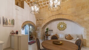 Uma sala de jantar com uma mesa de madeira redonda, cadeiras e um candelabro. Há um sofá com almofadas e uma peça decorativa na parede. Uma porta em arco dá acesso a outro espaço.