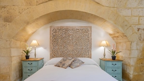 Um quarto com arco de pedra, uma cama com roupa de cama branca, duas mesas de cabeceira com luminárias e plantas em vasos, e um painel decorativo acima da cama.