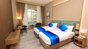 Habitación de hotel con dos camas, un escritorio, una silla, una lámpara y vistas a la ciudad.