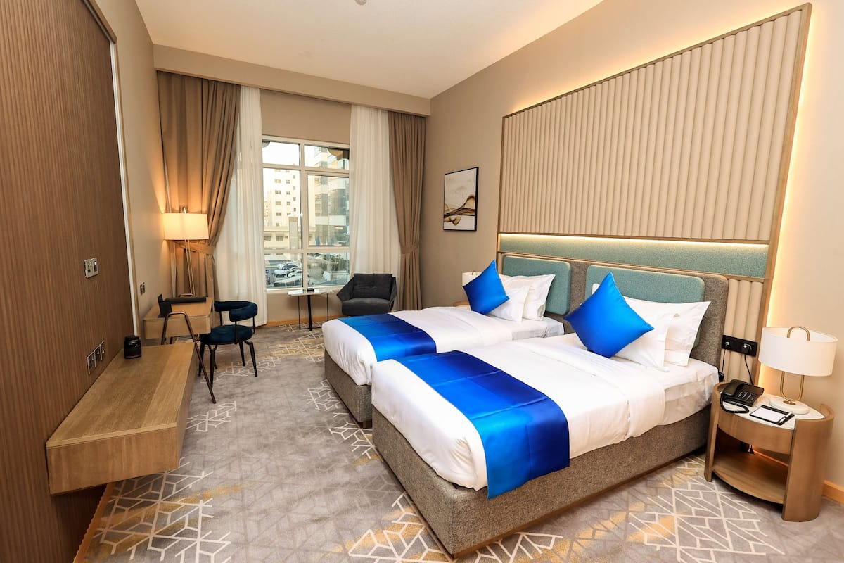 A hotel room with two beds, a desk, a chair, a lamp, and a view of the city.