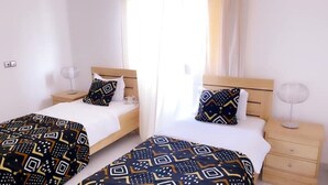 A hotel room with two single beds, each with patterned bedding, a bedside table with a lamp, and a window with sheer curtains. - 3-room apartment near the center. Air-conditioned and free WLan (Lomé)