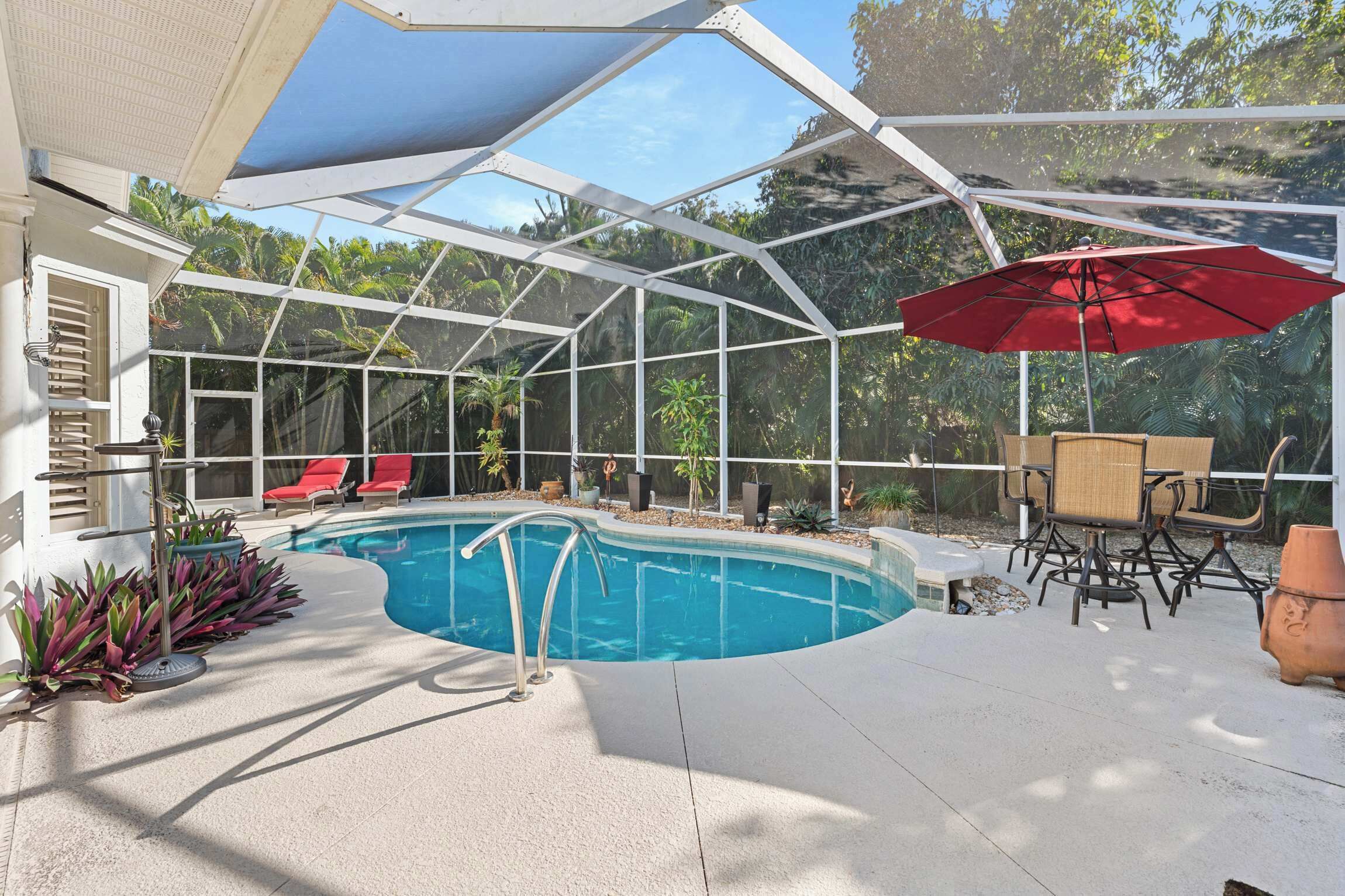 A covered outdoor swimming pool with a red umbrella, lounge chairs, and a patio table.