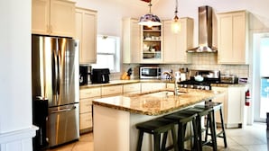 A modern kitchen with stainless steel appliances, a granite countertop, and a central island with bar stools. - Kokomo Cay - Luxurious Home Near the Beach, with Spectacular Ocean Views (Hope Town)