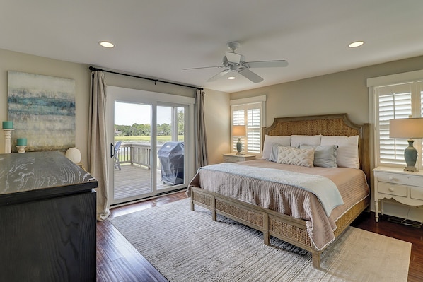 A bedroom with a large bed, a ceiling fan, a sliding glass door leading to a balcony, and a painting on the wall.