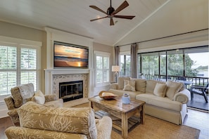 A spacious living room with a fireplace, a large TV mounted above it, and a view of the outdoors through large windows.