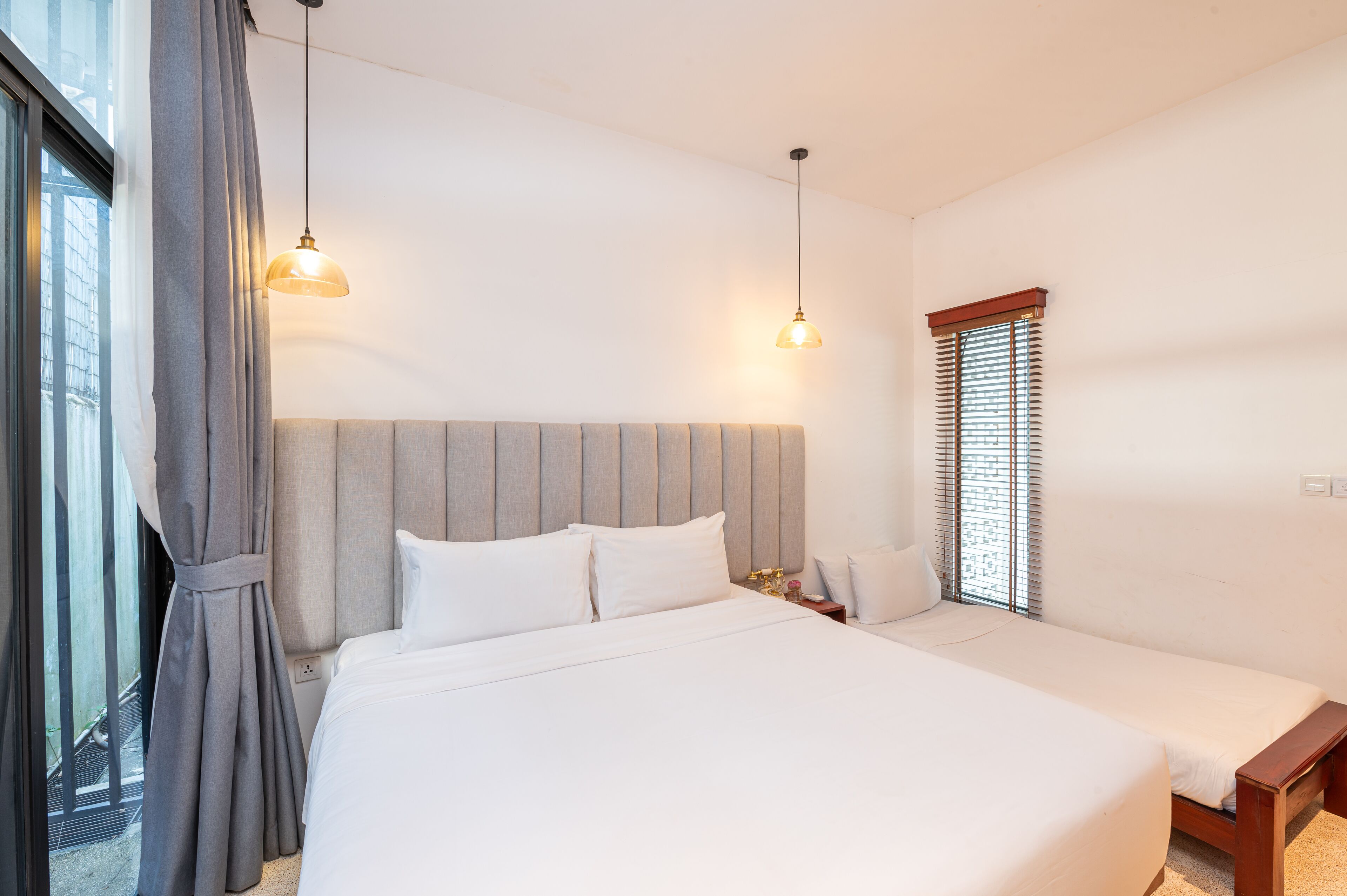 A hotel room with a large bed, a smaller bed, a nightstand, a lamp, and a window with blinds.