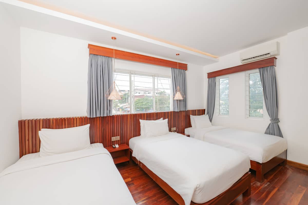 Une chambre d’hôtel avec trois lits, des têtes de lit en bois, un plancher en bois et de grandes fenêtres avec des rideaux gris.