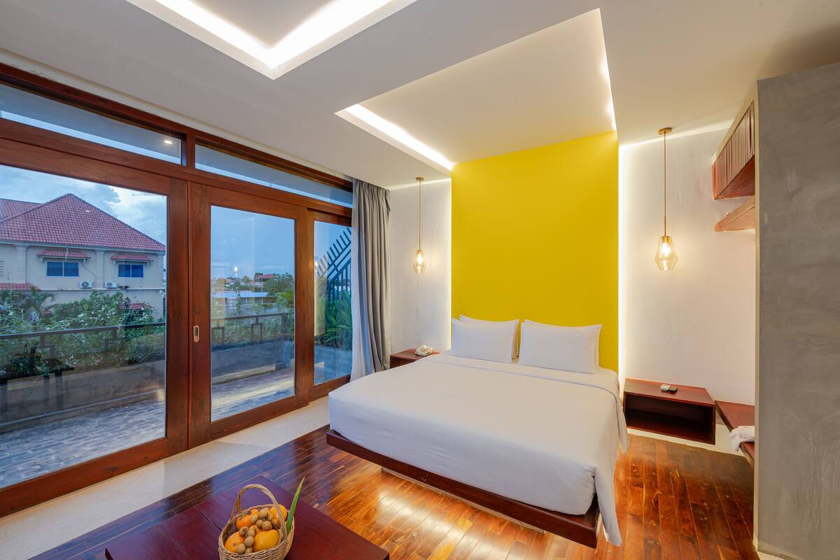 Une chambre d’hôtel moderne avec un grand lit, un plancher en bois, un mur d’accent jaune vif et une vue sur un quartier résidentiel grâce à de grandes portes coulissantes.