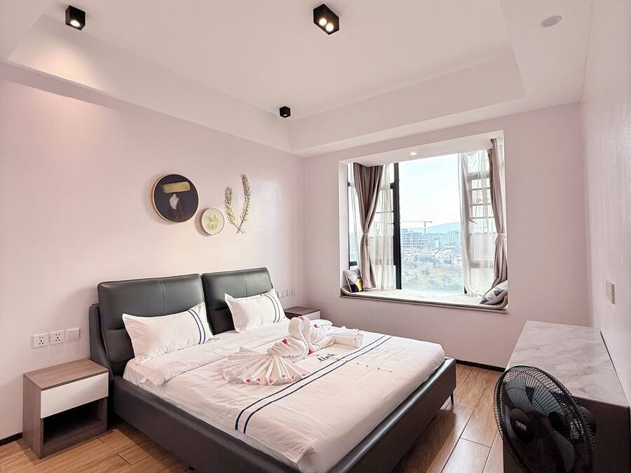 Une chambre moderne avec un grand lit, une table de chevet, un ventilateur sur pied et une fenêtre donnant sur la ville.